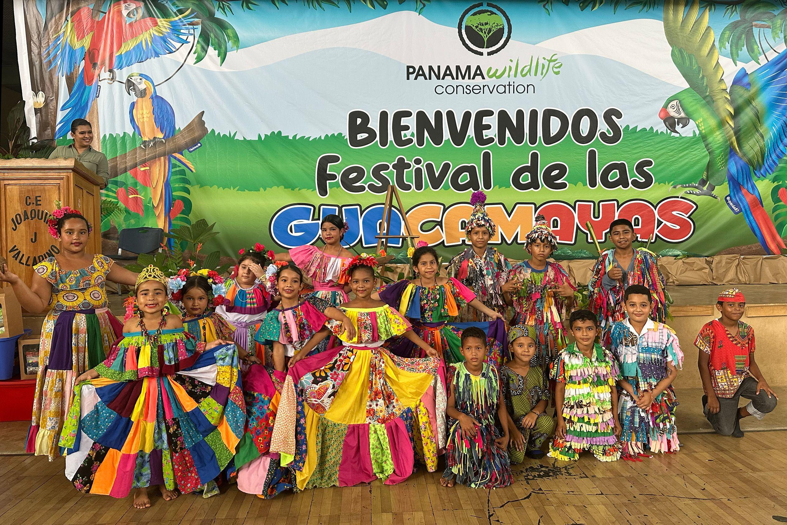 Children in colorful traditional outfits pose on a stage during Panama Wildlife Conservation’s Festival de las Guacamayas, in front of a welcome banner illustrated with macaws
