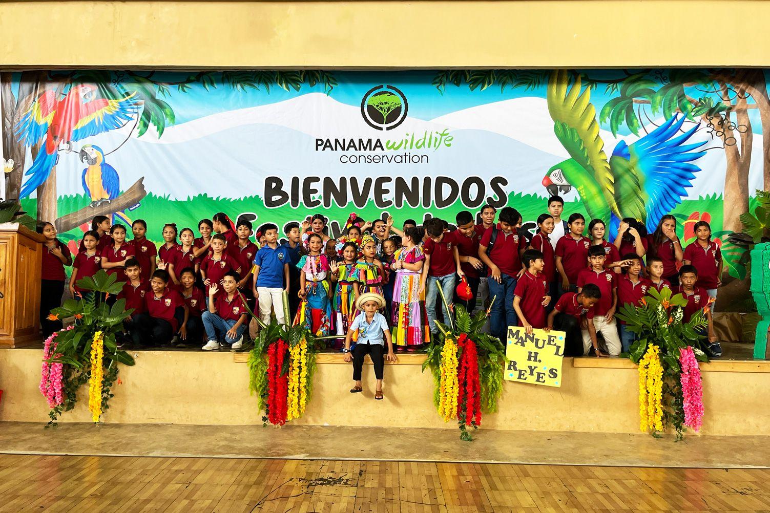 Group of schoolchildren on stage at Panama Wildlife Conservation’s Festival de las Guacamayas, with some wearing traditional colorful clothing in front of a macaw-themed conservation banner.